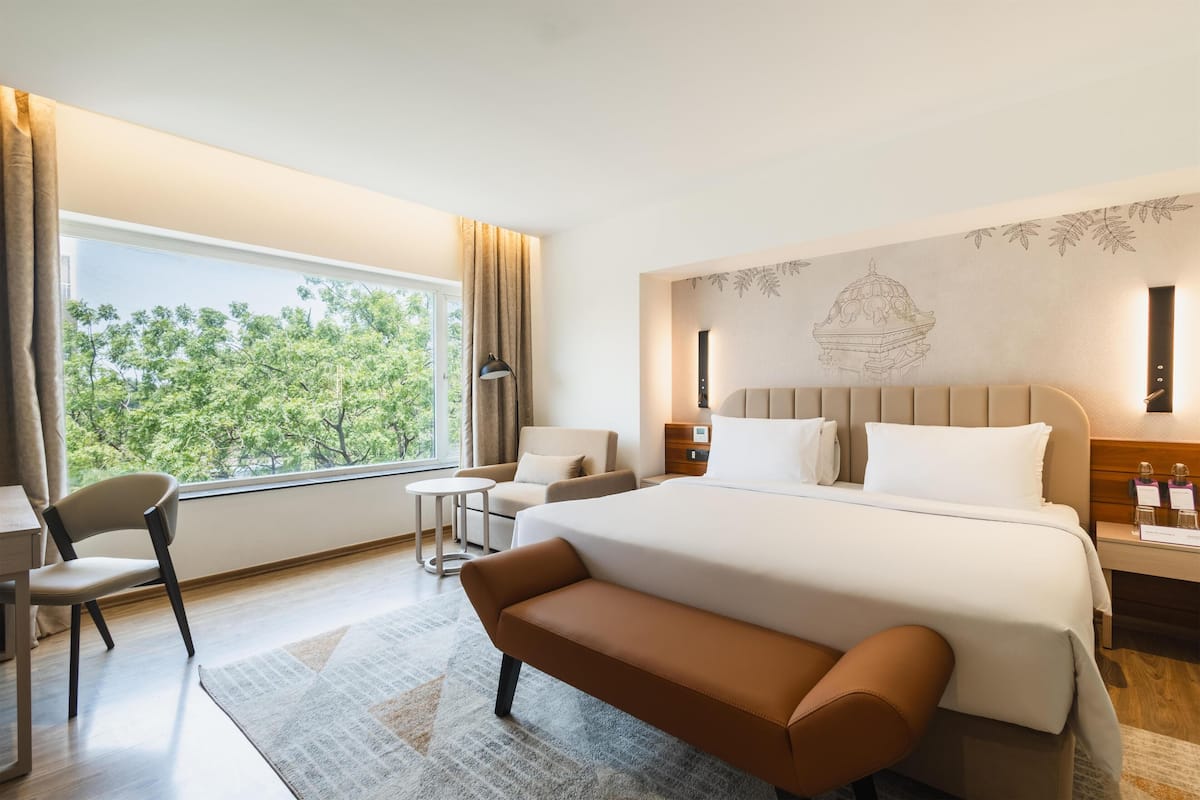 Quarto de hotel com uma cama grande, uma escrivaninha, uma cadeira, uma mesa pequena e vista para as árvores pela janela.