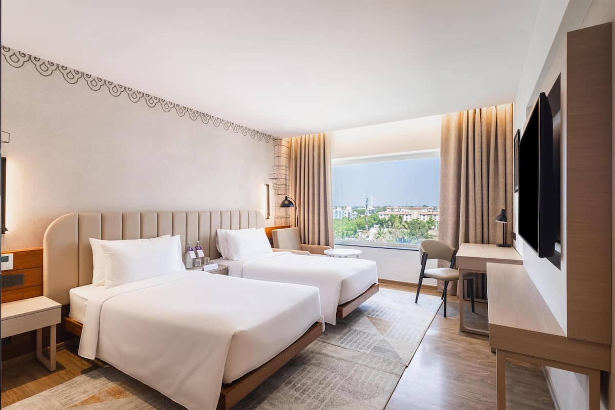 Quarto de hotel com duas camas, uma escrivaninha, uma cadeira, televisão e janela com vista para a cidade.