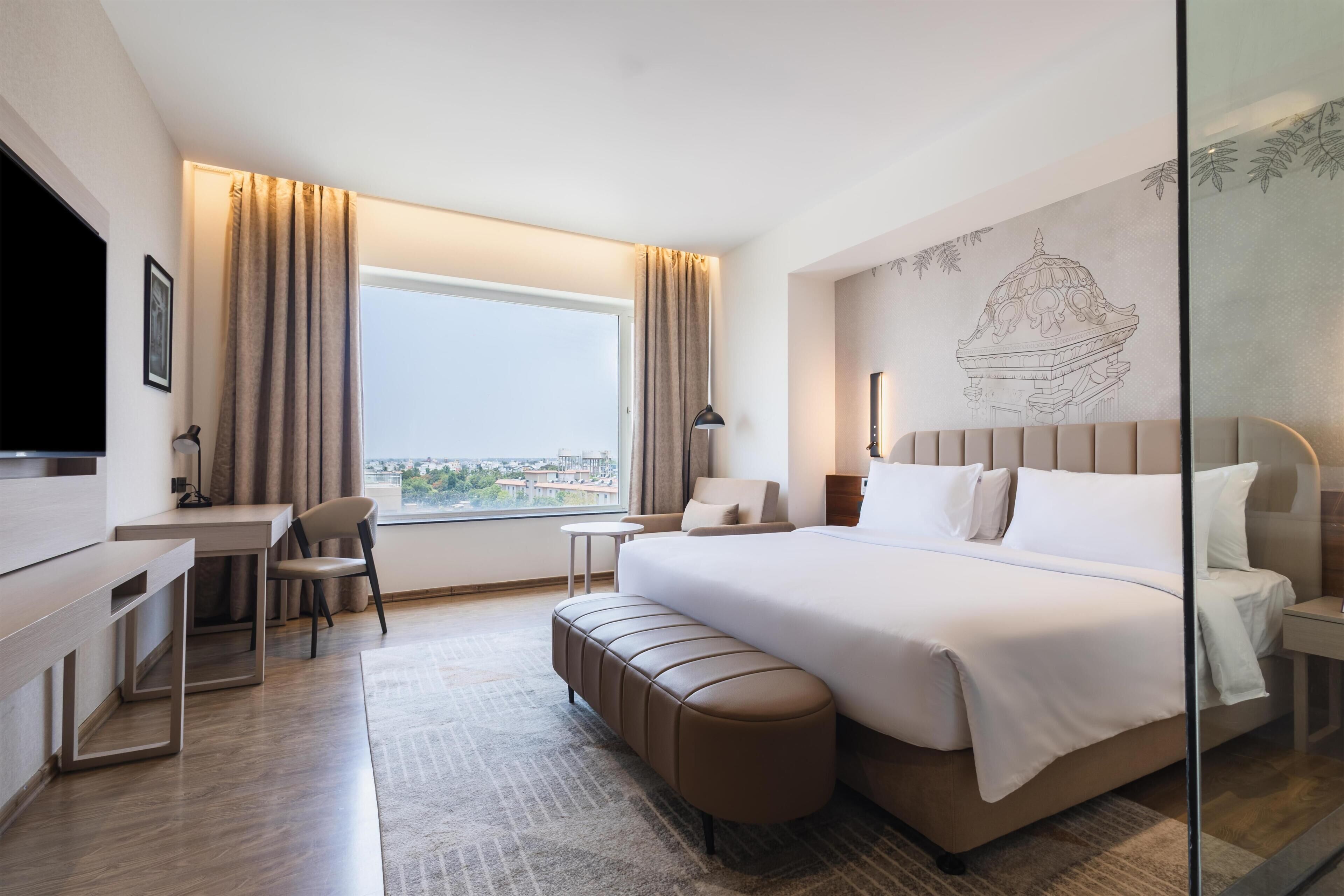 A hotel room with a bed, a desk, a chair, a bench, a TV, and a large window with curtains.