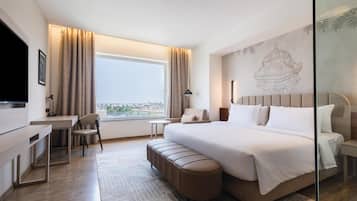 A hotel room with a bed, a desk, a chair, a bench, a TV, and a large window with curtains.