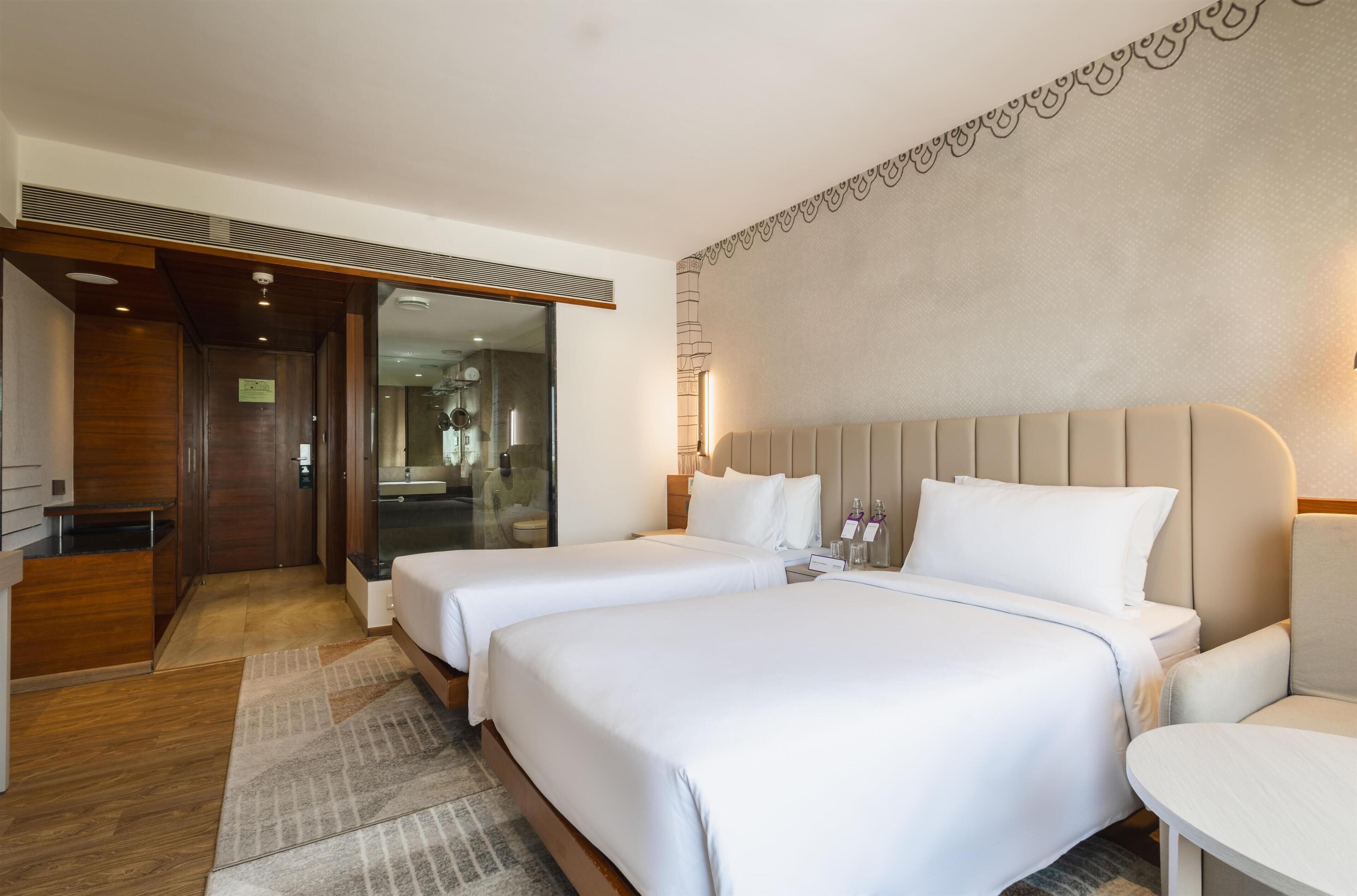 A hotel room with two beds, a wooden wardrobe, a bathroom with a glass door, and a patterned wall.