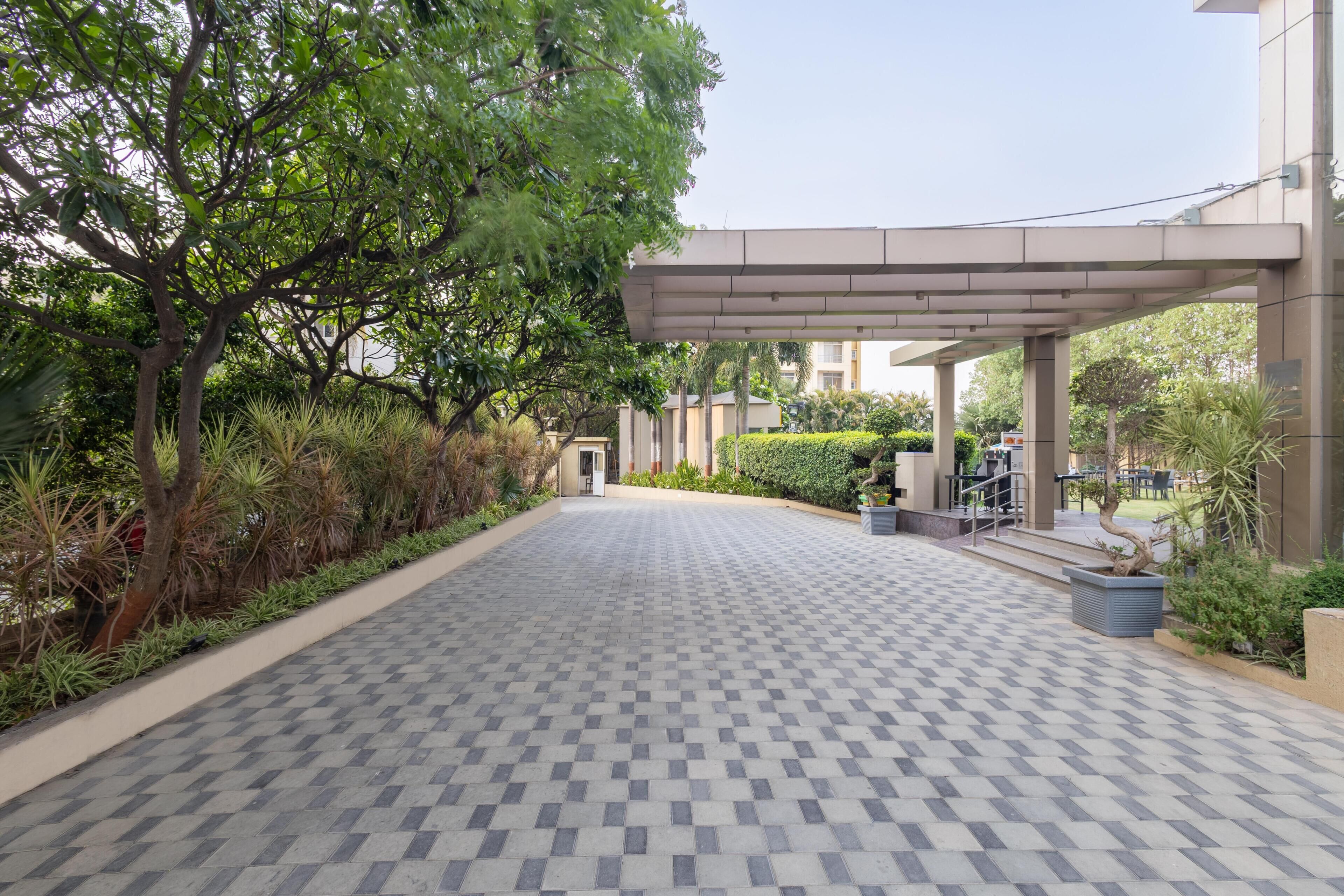 A paved walkway leading to a building with a covered entrance, surrounded by greenery and a tiled floor.