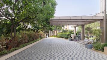 A paved walkway leading to a building with a covered entrance, surrounded by greenery and a tiled floor.