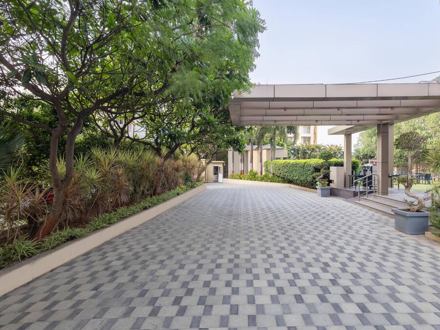 A paved walkway leading to a building with a covered entrance, surrounded by greenery and a tiled floor.