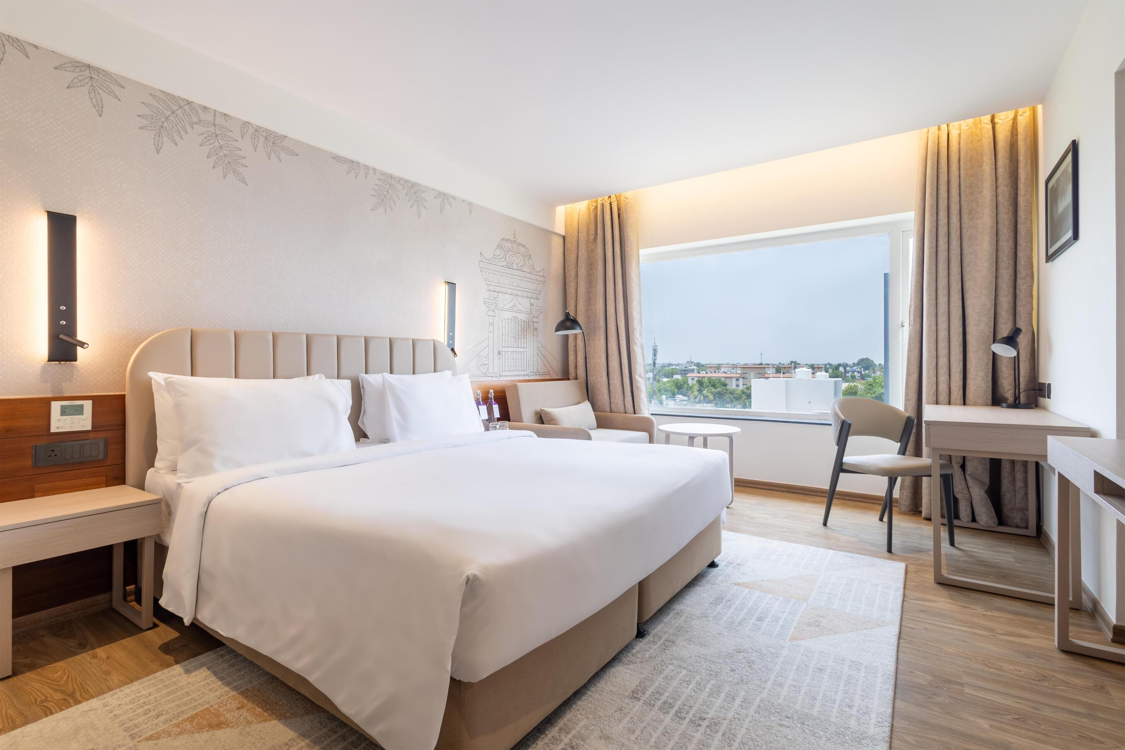 Habitación de hotel con una cama grande, mesitas de noche, un escritorio, una silla y vistas a la ciudad.