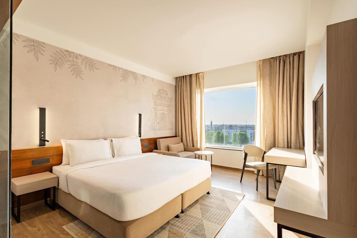 Quarto de hotel com cama grande, escrivaninha, cadeira, mesinha de centro e vista da cidade pela janela.
