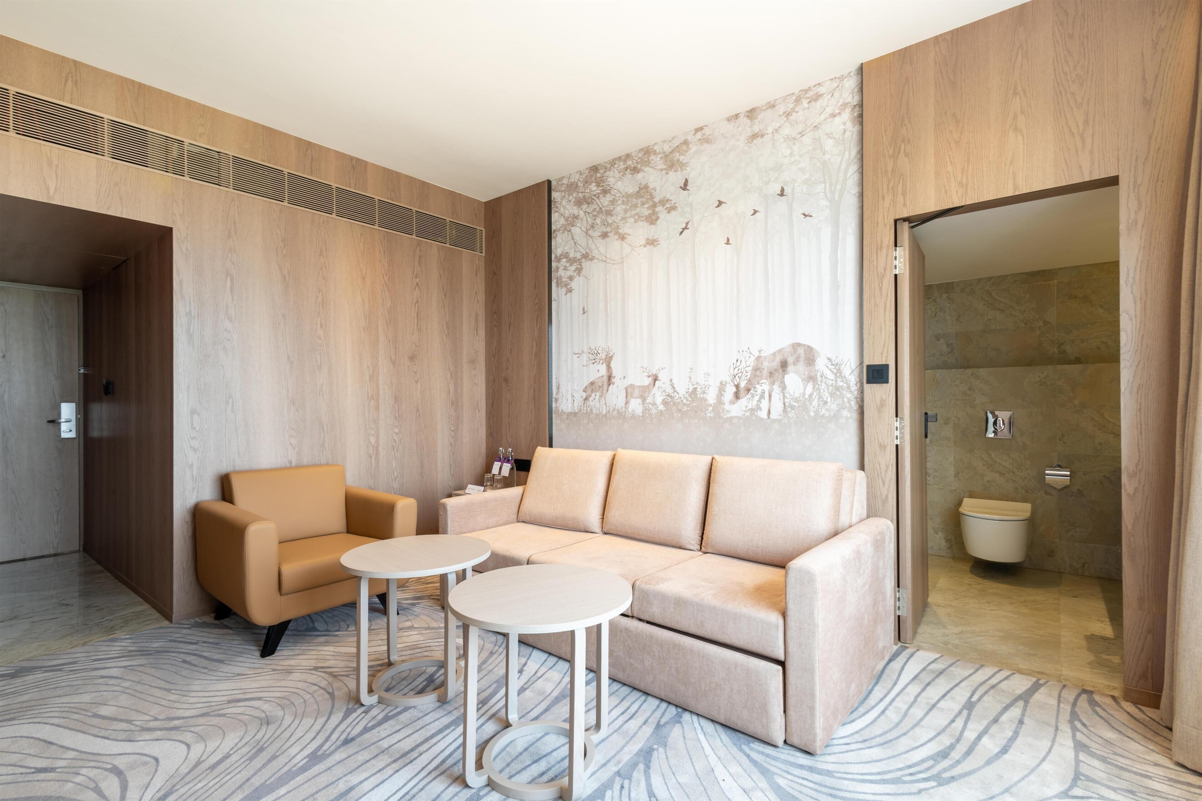 Una habitación de hotel moderna con un sofá color beige, dos mesas redondas, un mural con ciervos y árboles, y un baño visible a través de una puerta abierta.