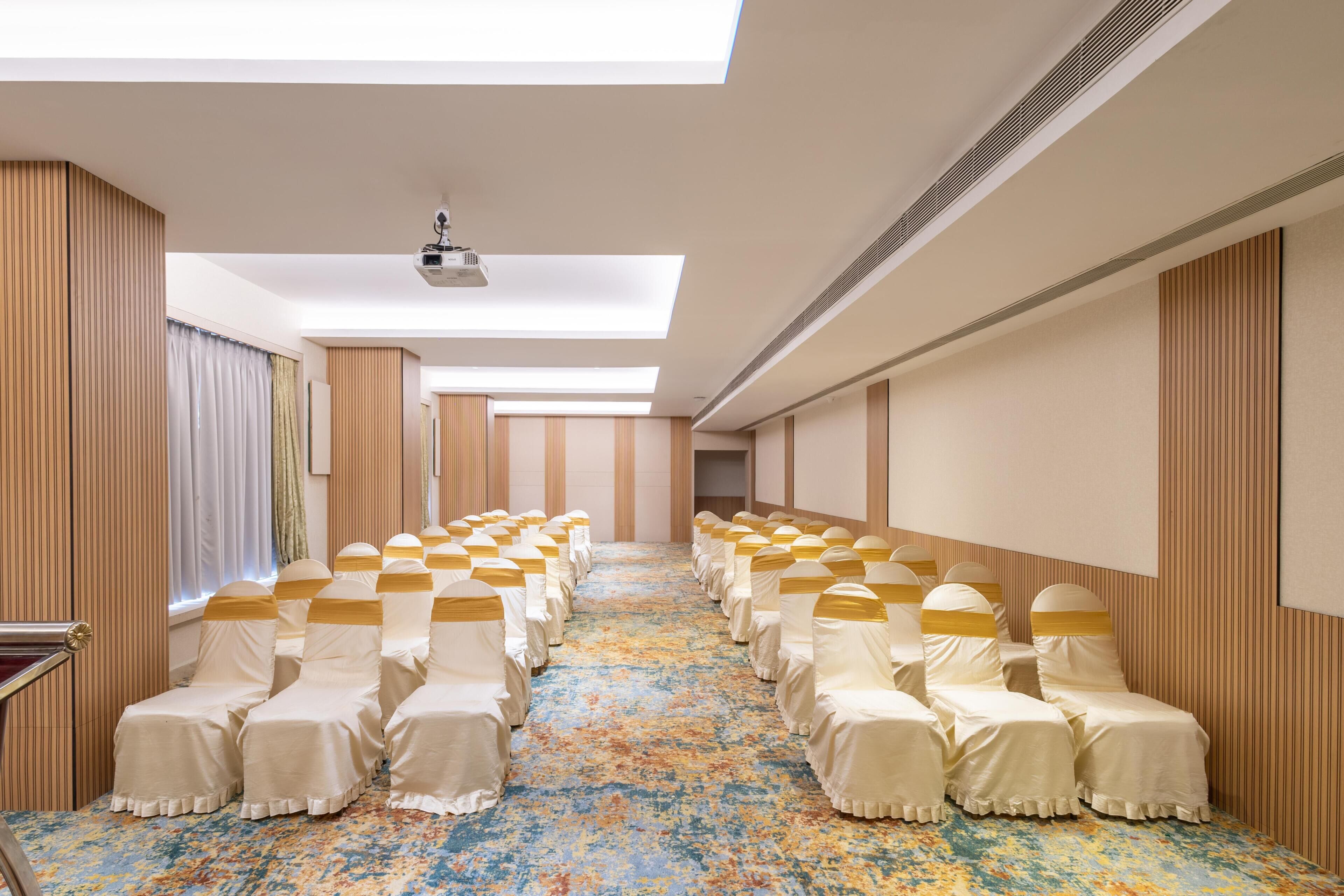 Un salón de hotel con una hilera larga de sillas cubiertas con manteles blancos y fajas doradas, una alfombra estampada y un techo con tragaluces.