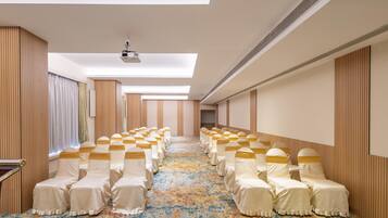 Un salón de hotel con una hilera larga de sillas cubiertas con manteles blancos y fajas doradas, una alfombra estampada y un techo con tragaluces.