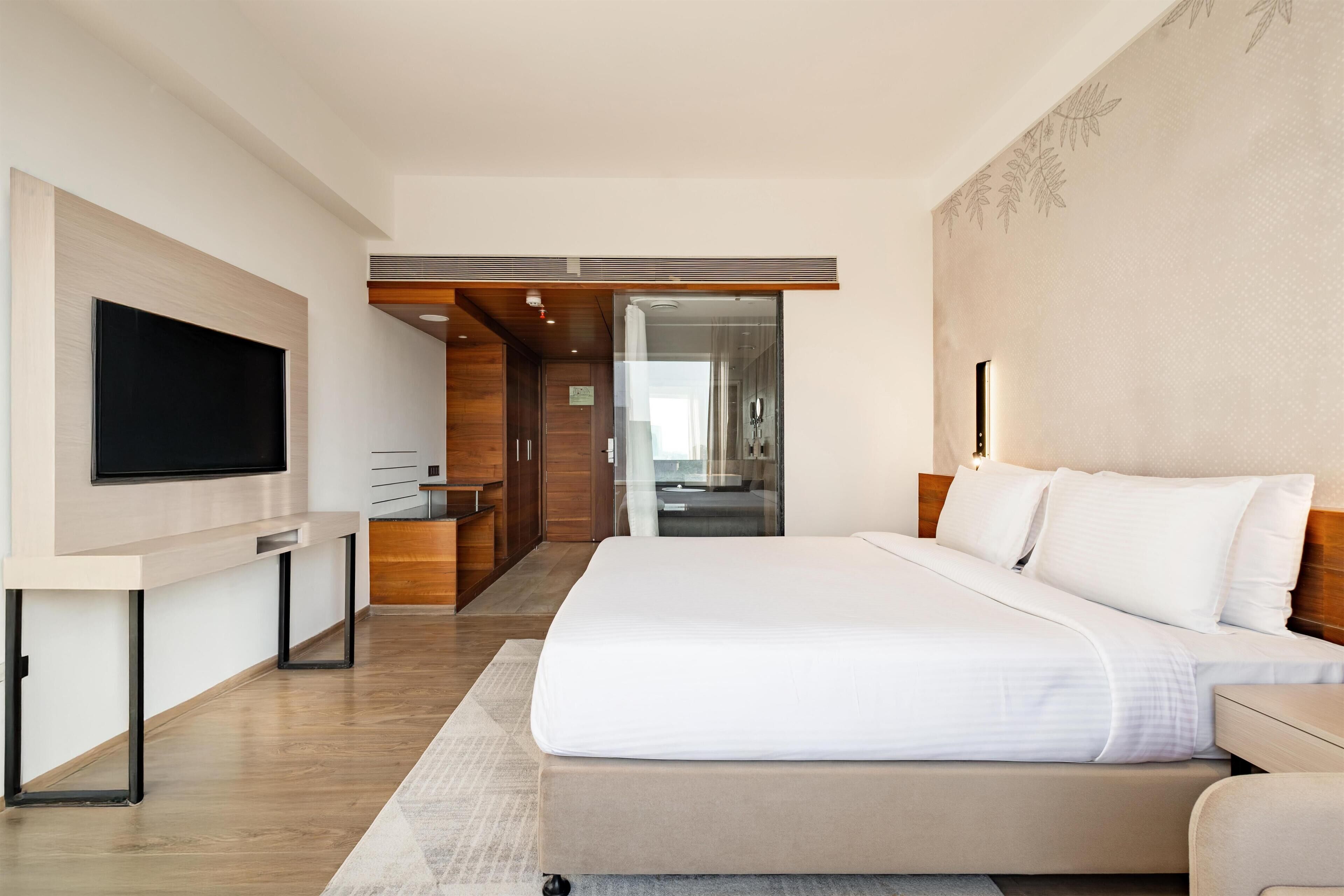 Habitació d'hotel moderna amb un llit gran, televisió de pantalla plana penjada a la paret, escriptori amb cadira i porta de vidre que dona a un bany.