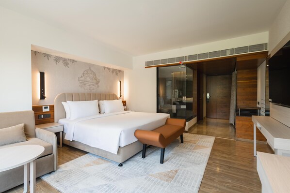 A modern hotel room with a large bed, a seating area, a TV, and a bathroom with a glass door.