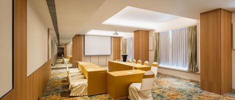 Un salón de conferencias con una larga mesa, sillas, una pantalla de proyector y un amplio ventanal con cortinas.
