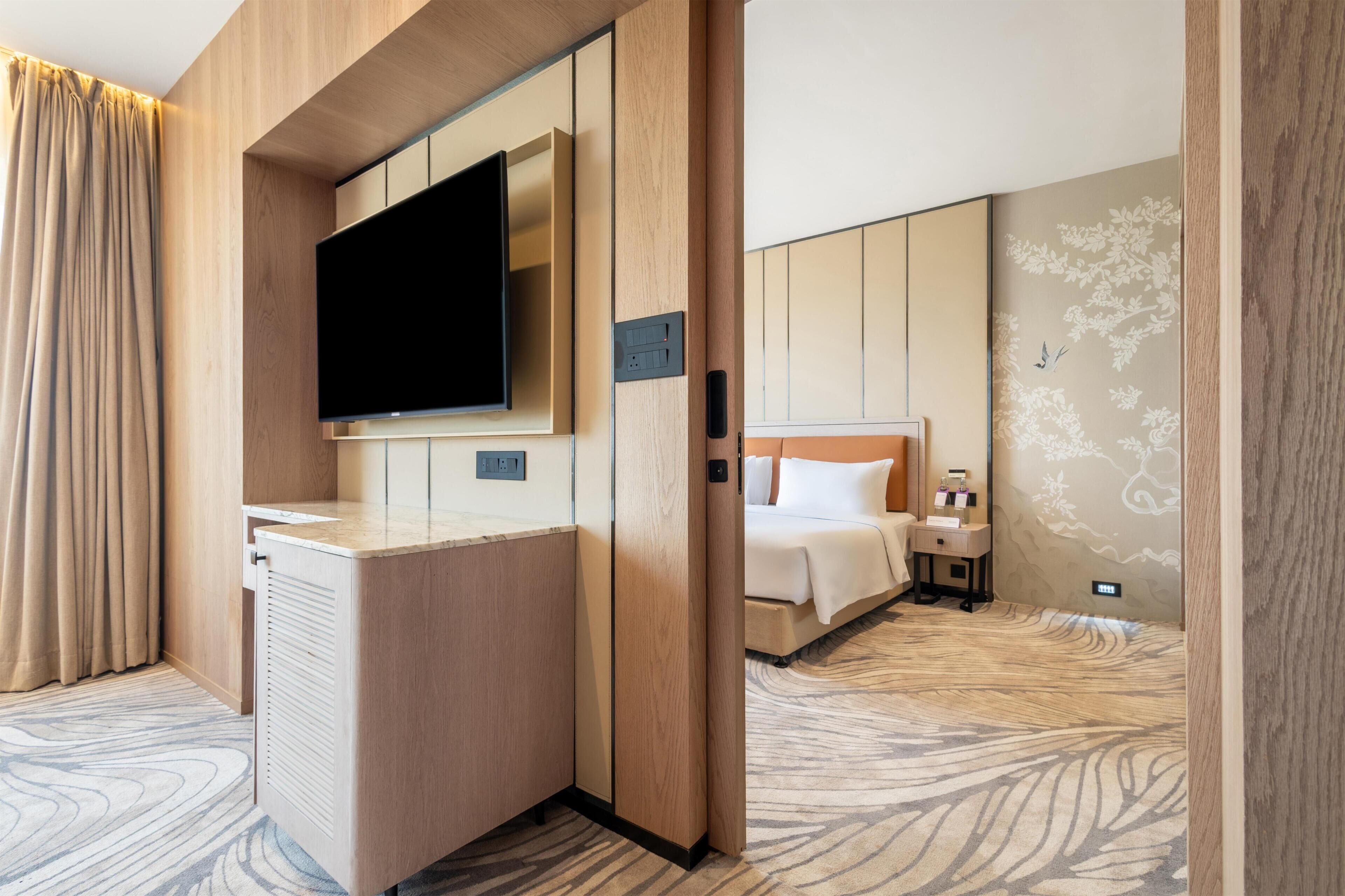 A hotel room with a bed, a TV mounted on a wooden panel, a bedside table, and a patterned carpet.