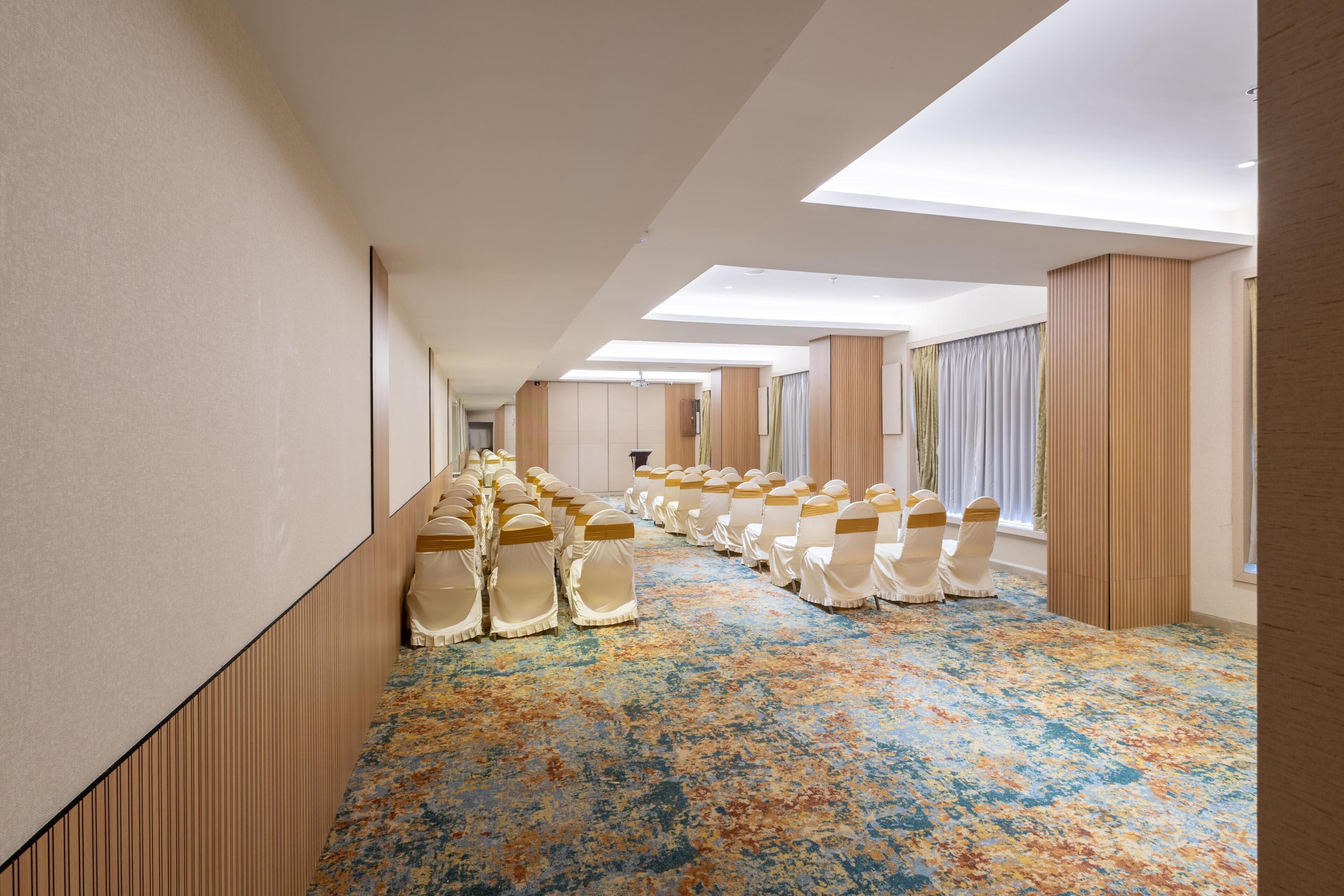 Un pasillo de hotel con filas de sillas, una alfombra estampada y un techo alto con tragaluces.
