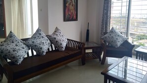 A living room with a wooden sofa, floral cushions, a glass coffee table, and a painting on the wall. - Kharadi EON IT NP Hospitality (Pune)