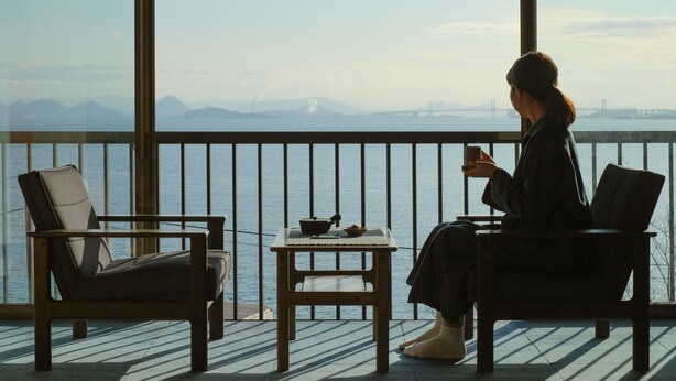 A person sitting by a window overlooking a body of water with mountains and a bridge in the distance. - DENIM HOSTEL float (Kurashiki)