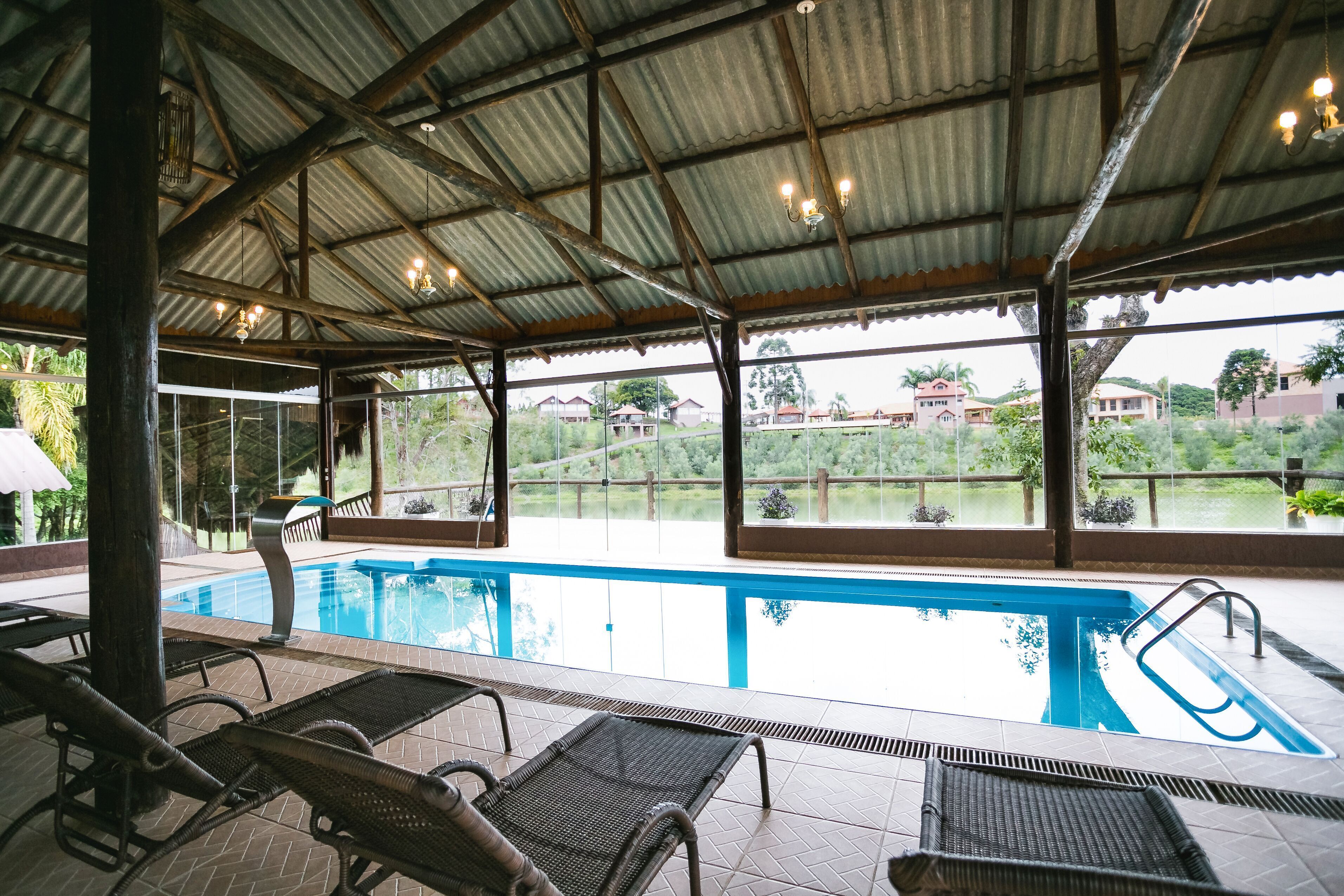 An indoor swimming pool with clear glass walls, lounge chairs, and a view of the surrounding landscape.
