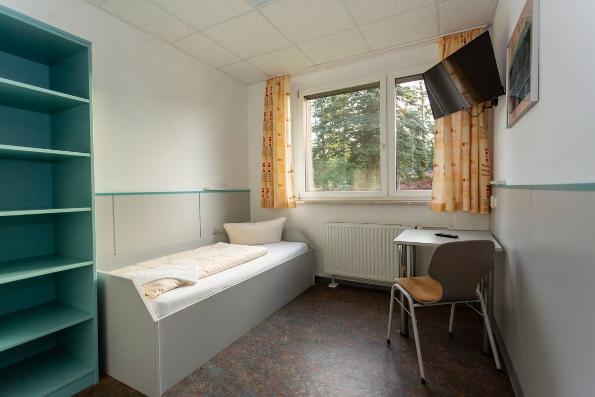 A small, single-bed room with a window, a desk, and a chair.