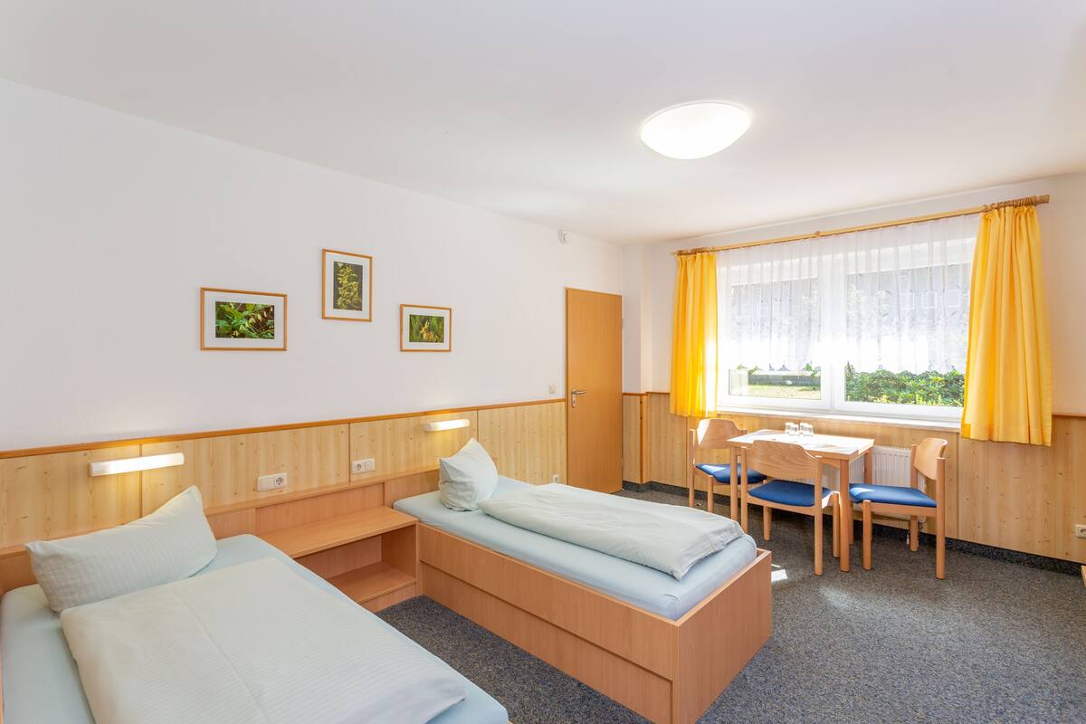A hotel room with two beds, a desk, and chairs. There are three framed pictures on the wall.