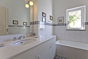 A bathroom with a white sink, vanity, and bathtub. There are framed pictures on the wall, a window with a view of greenery, and a tiled backsplash.