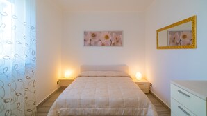A bedroom with a bed, bedside lamps, a nightstand, a dresser, a mirror, and a framed floral artwork on the wall. - Appartamento Molo B (Portoferraio)