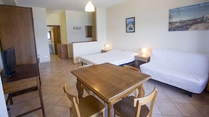 Une petite pièce bien éclairée, avec une table et des chaises en bois, un canapé blanc, un lit et un téléviseur posé sur un bureau.