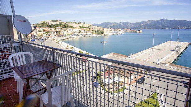 Un balcon avec une table et des chaises, donnant sur une ville côtière avec un port de plaisance et des montagnes en arrière-plan.