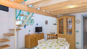 A room with a wooden ceiling, a dining table with a tablecloth, a television on a wooden cabinet, a fish wall decoration, and a cabinet with glass doors. - Appartamento Poggio Al Sole (Capoliveri)