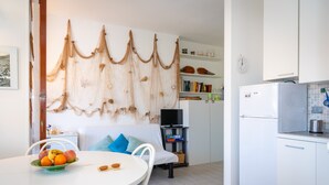 A modern kitchen with white cabinets, a built-in oven, and a refrigerator. There is a dining area with a table set for two, a television, and a bookshelf. - Appartamento Porto Mediceo (Portoferraio)