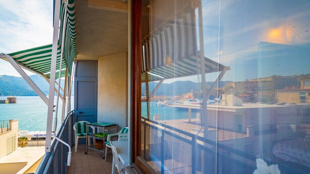 A balcony with a view of the sea and mountains, a table with chairs, and a window with a flower. - Appartamento Porto Mediceo (Portoferraio)
