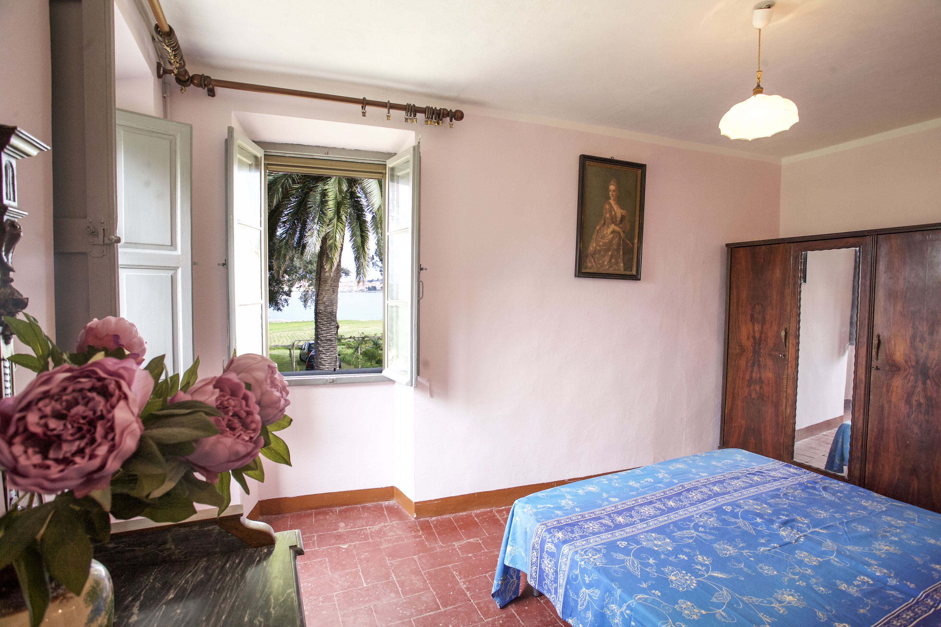 A bedroom with a bed, a window with a view of palm trees, a wooden wardrobe, and a painting on the wall.