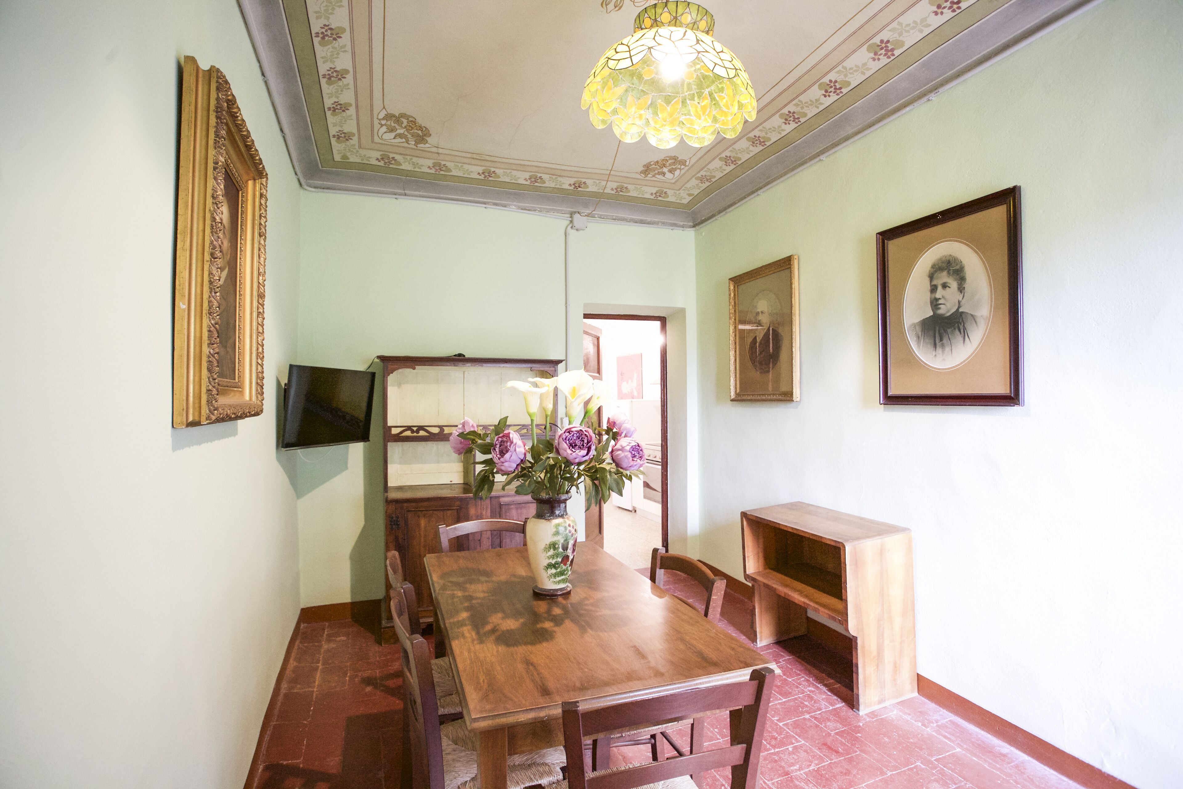 Un comedor con mesa de madera, sillas, un florero con flores, cuadros en la pared y una araña.