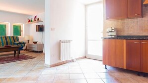 Una sala de estar con sofá, una mesa de centro y un televisor sobre una estantería. Hay una zona de cocina con armarios de madera y piso de baldosas.