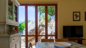 A dining area with a table set for two, a television, and a view of a rocky landscape through large glass doors. - Villino Forno (Portoferraio)