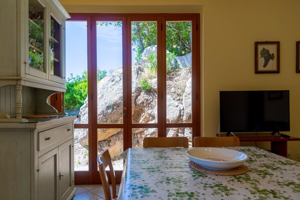 Un coin repas avec une table dressée pour deux personnes, un téléviseur et une vue sur un paysage rocheux, visible à travers de grandes portes vitrées.