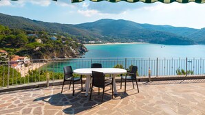 A terrace with a table and chairs overlooking a coastal landscape with a beach and mountains. - Villino Forno (Portoferraio)