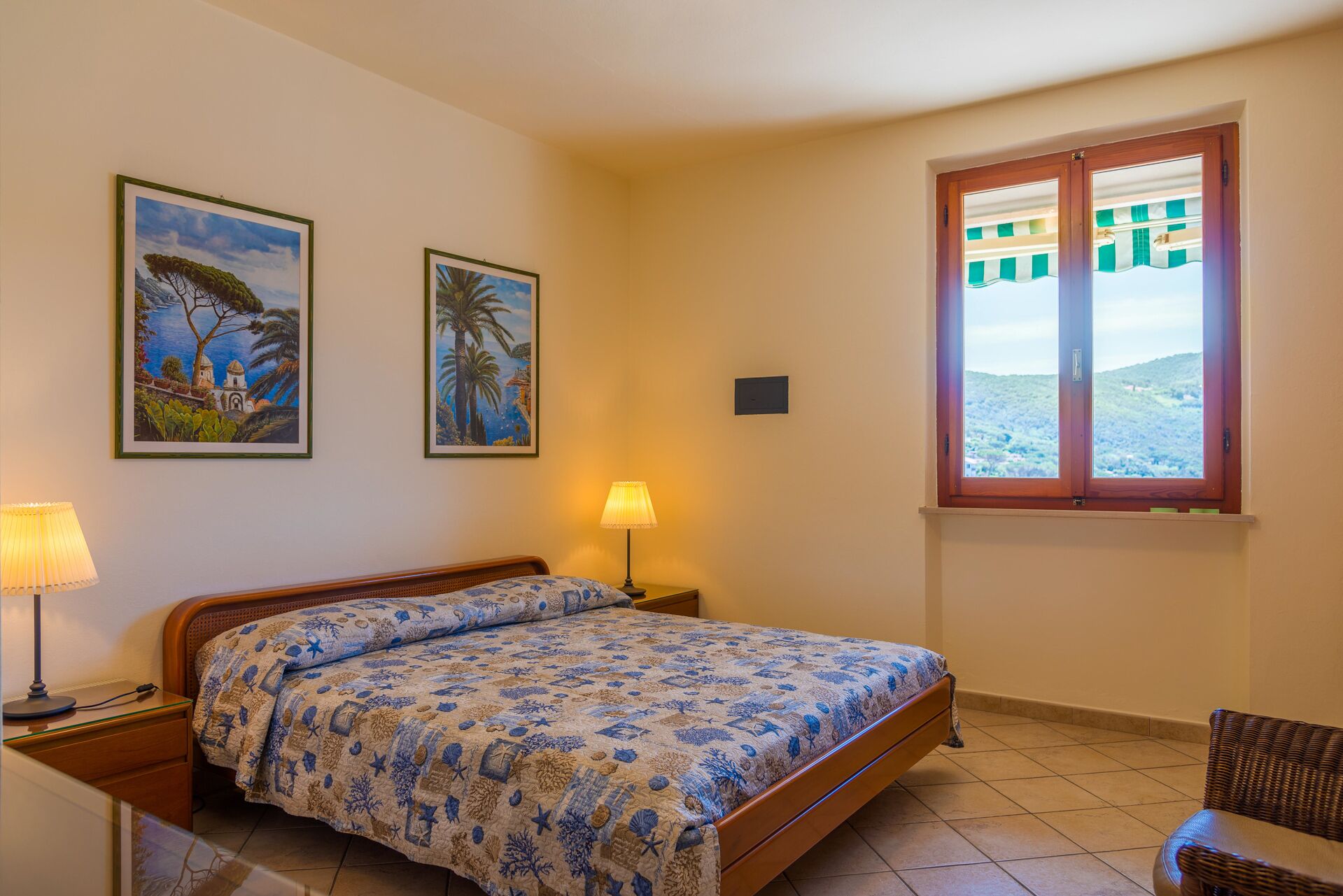 Quarto com cama, duas luminárias na parede, duas pinturas emolduradas, uma janela com vista e uma cadeira.