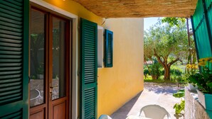 A covered outdoor patio with a table and chairs, a door with shutters, and a view of greenery. - Villino La Fontanella (Portoferraio)