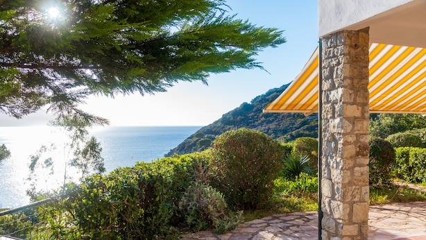 A stone pathway leading to a building with a yellow-striped awning, surrounded by lush greenery and overlooking a coastal landscape. - Appartamenti Panorama (Portoferraio)