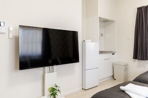 A modern hotel room with a flat-screen TV mounted on the wall, a compact kitchenette with a refrigerator, and a bed with a dark-colored blanket.