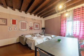 A room with a bed, a table, and a lamp. There are framed pictures on the wall.