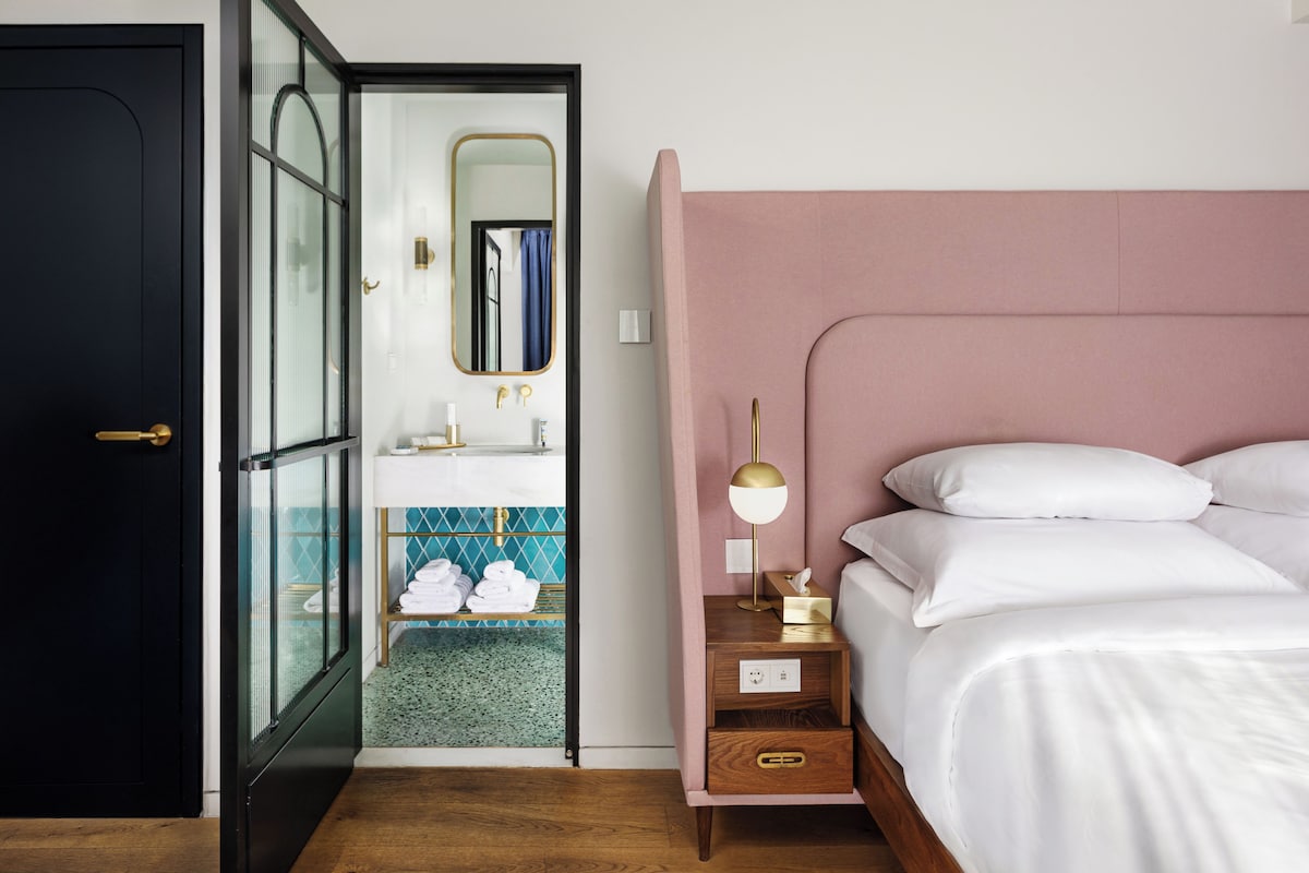 A bedroom with a bed, bedside table, lamp, and a bathroom with a sink and mirror.