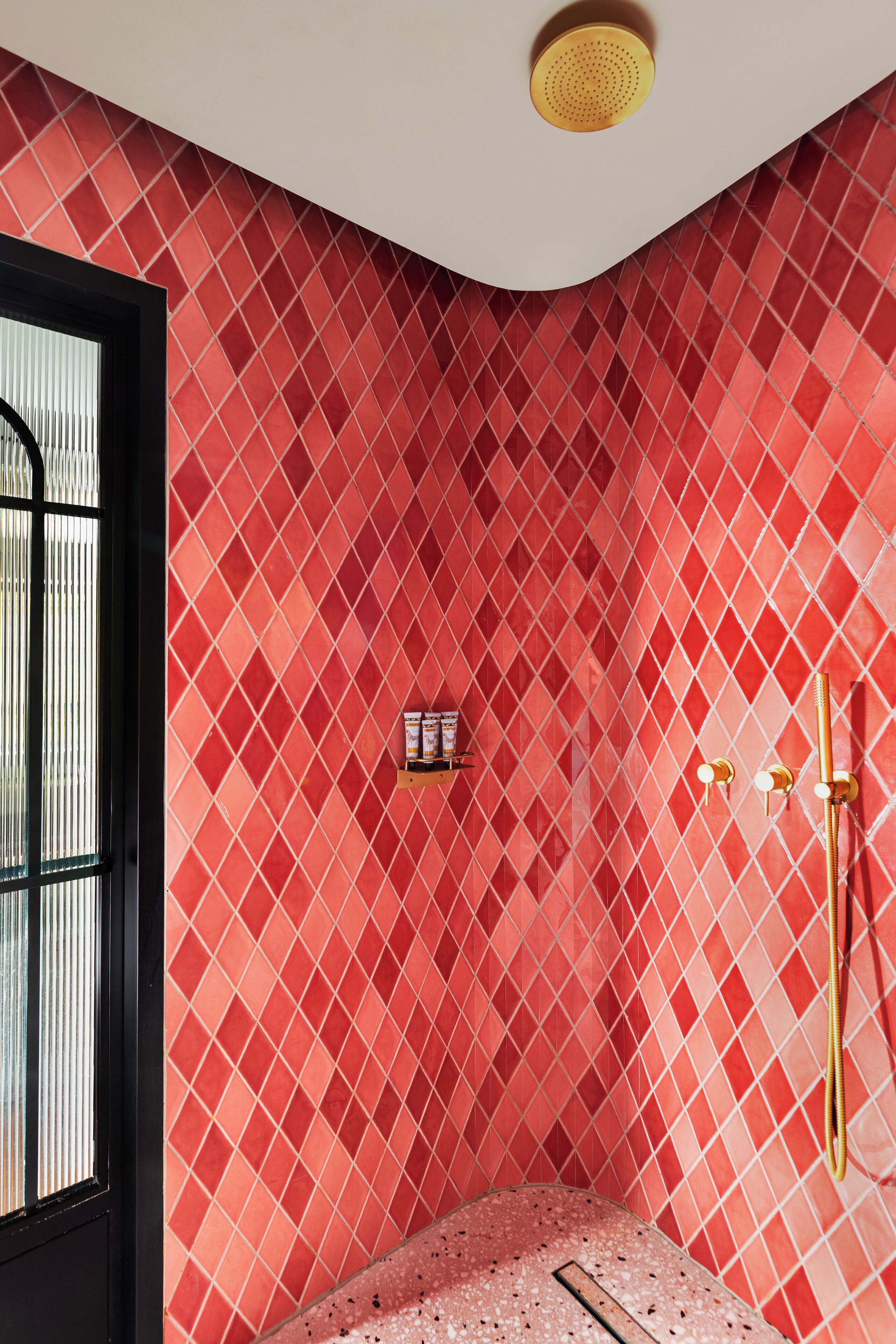 Un baño con pared de baldosas rojas con diseño de rombos, ducha con detalles dorados y ventana con persianas verticales.