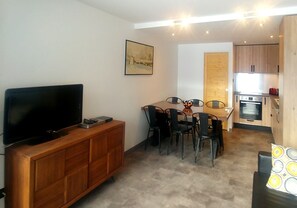 A compact living space with a TV on a wooden cabinet, a dining table with chairs, a kitchen area with wooden cabinets, and a painting on the wall.
