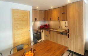 A compact kitchen with wooden cabinets, a built-in oven, a sink, and a dining table with a bowl of oranges.