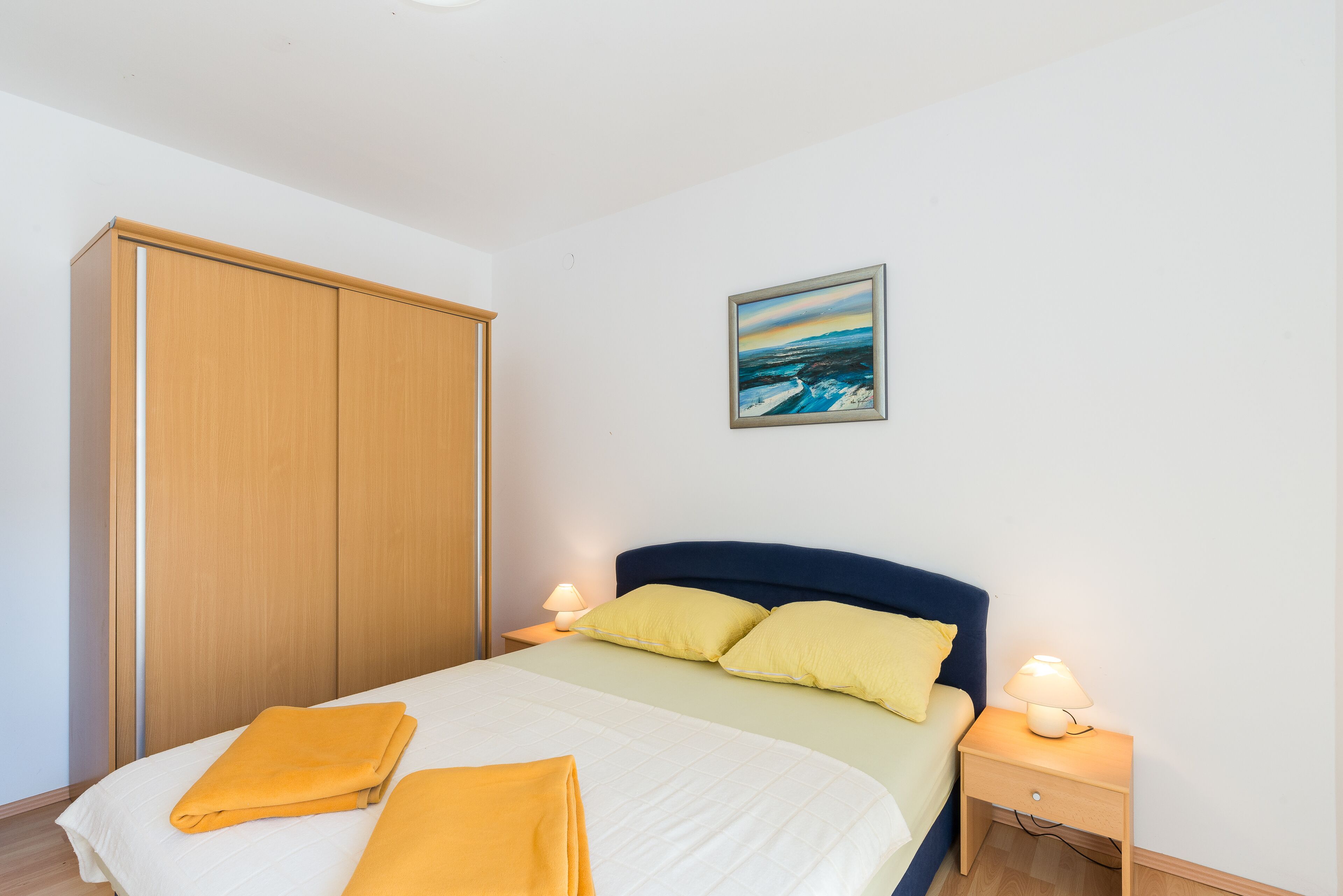 A bedroom with a bed, two bedside lamps, a nightstand, a wooden wardrobe, and a painting on the wall.