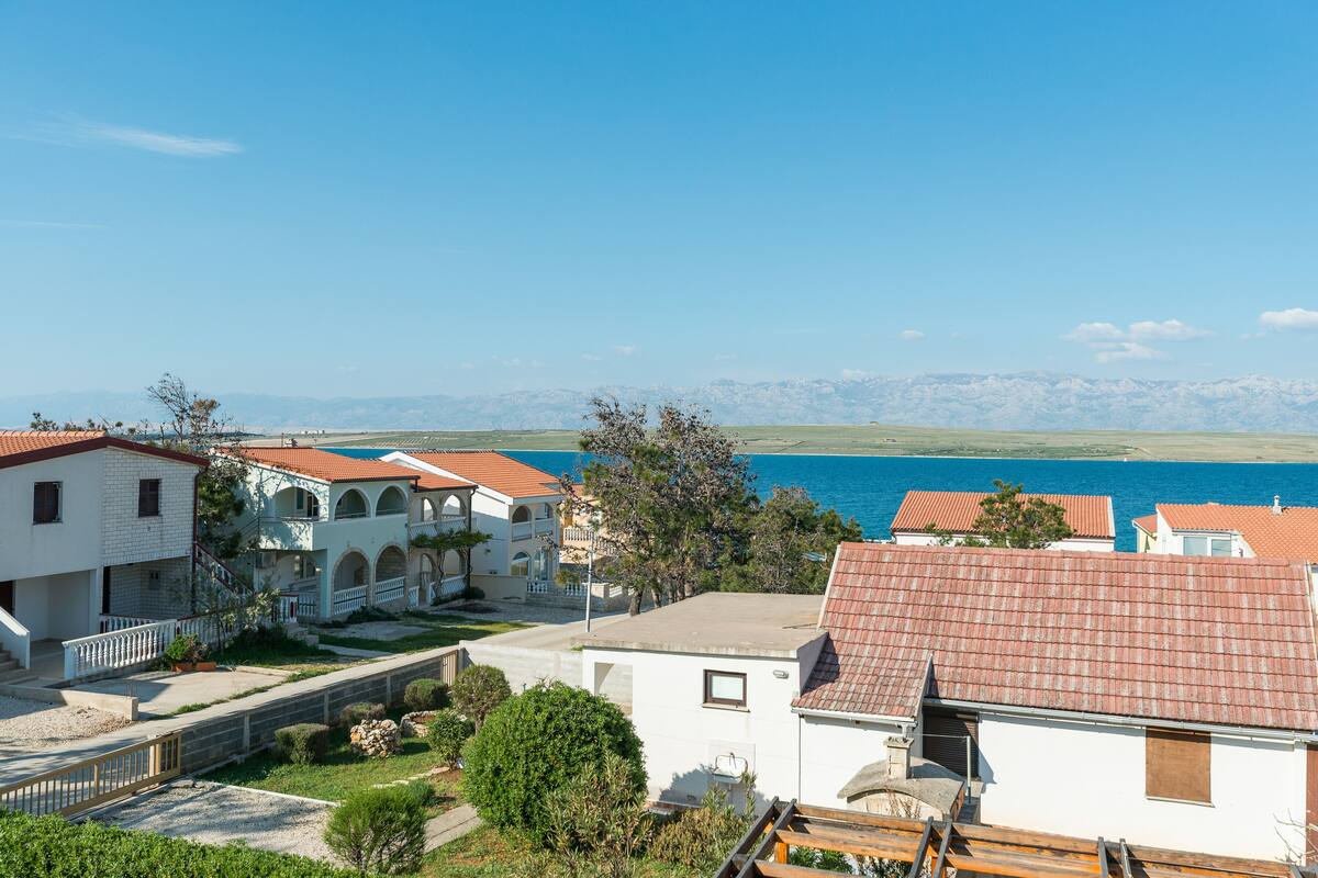 Ein Küstenwohngebiet mit weißen Gebäuden, roten Ziegeldächern und freiem Blick auf das Meer und die dahinterliegenden Berge.