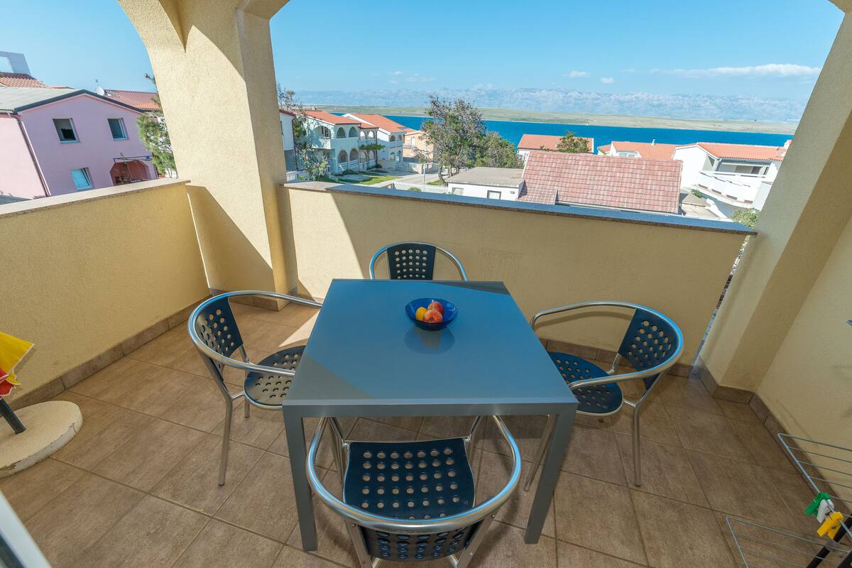 Ein Balkon mit Tisch und Stühlen, einer Obstschale und Blick auf Gebäude sowie ein Gewässer.