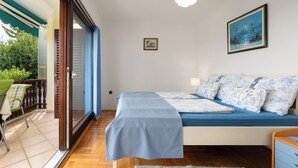 A bedroom with a bed, a small table, and a chair. There is a balcony with a view of greenery. - Funtana Apartments Budinjaš  (FUNTANA)