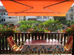 Ein Außenbereich mit Tisch und Stühlen unter einer orangefarbenen Markise, umgeben von üppiger Bepflanzung und roten Blüten.
